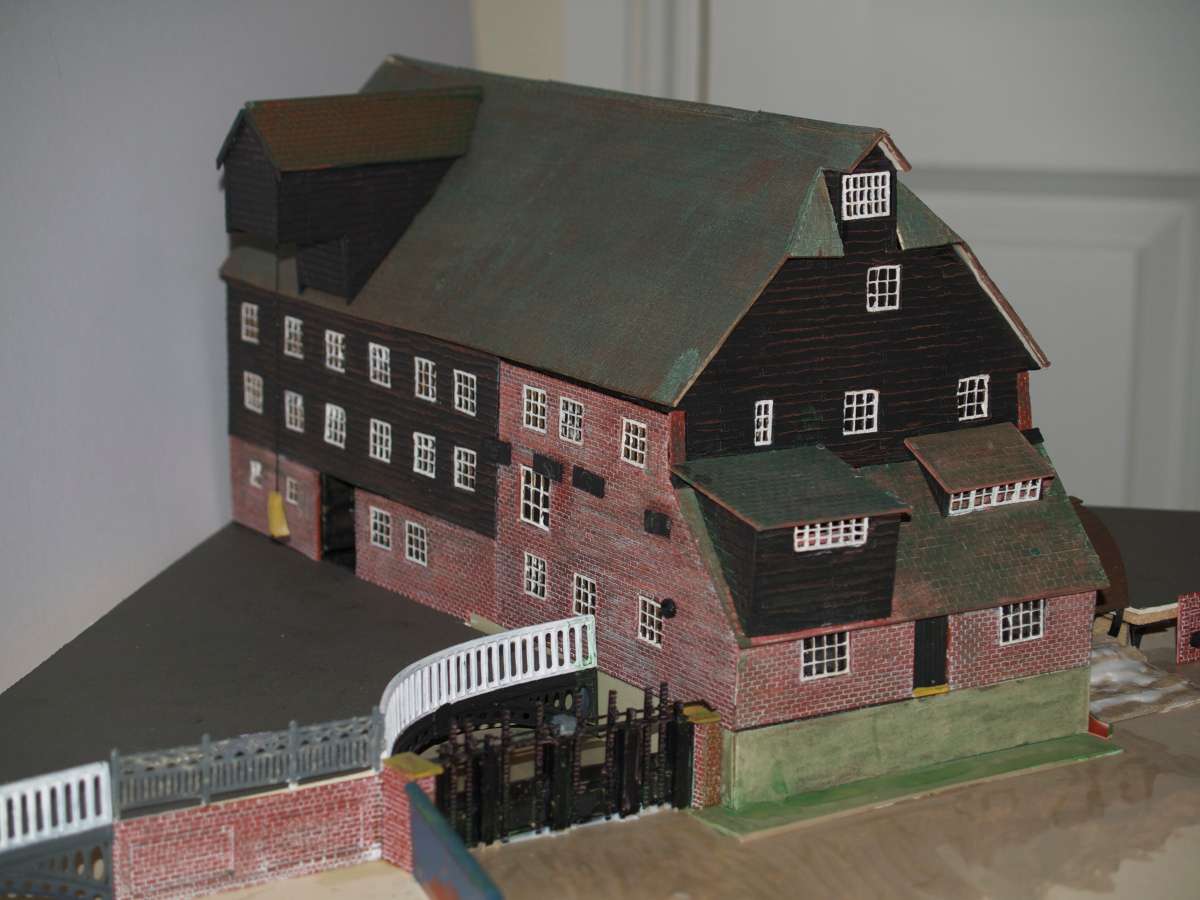 This is an OO scale model of Houghton Mill on the River Great Ouse at Houghton, near Huntingdon, Cambridgeshire. The mill is owned and operated as a working flour mill by the National Trust. I volunteer at the mill mainly in the construction and repair of interactive displays. We decided that during this closed season we would build a new attraction telling of the passage of grain from seed to table. I have obtained some 5 foot diameter circular tables which will be used, one is as base, the other is just the top which will rotate on the base. The display will be in quarters showing :- Ploughing and Sowing with Winter, Spring and Harvest, The Mill and finally a Bakers Shop with Tea Room. It is intended that the visitors will turn the top to see the various parts of the model through a window. The mill has been constructed on a mount board armature using proprietary plastic walling and LCut roofing and windows, the windows were a special order made to size, a total of 62 were made for me. I used a set of photocopied plans with a large number of photographs to get the proportions correct and incorporate as many details as possible. In order to get a good effect I have had to use a certain amount of license with some details but I hope that the overall effect will be pleasing to our visitors. We expect to have it on display when the Mill reopens to the public on 26th March 2015.